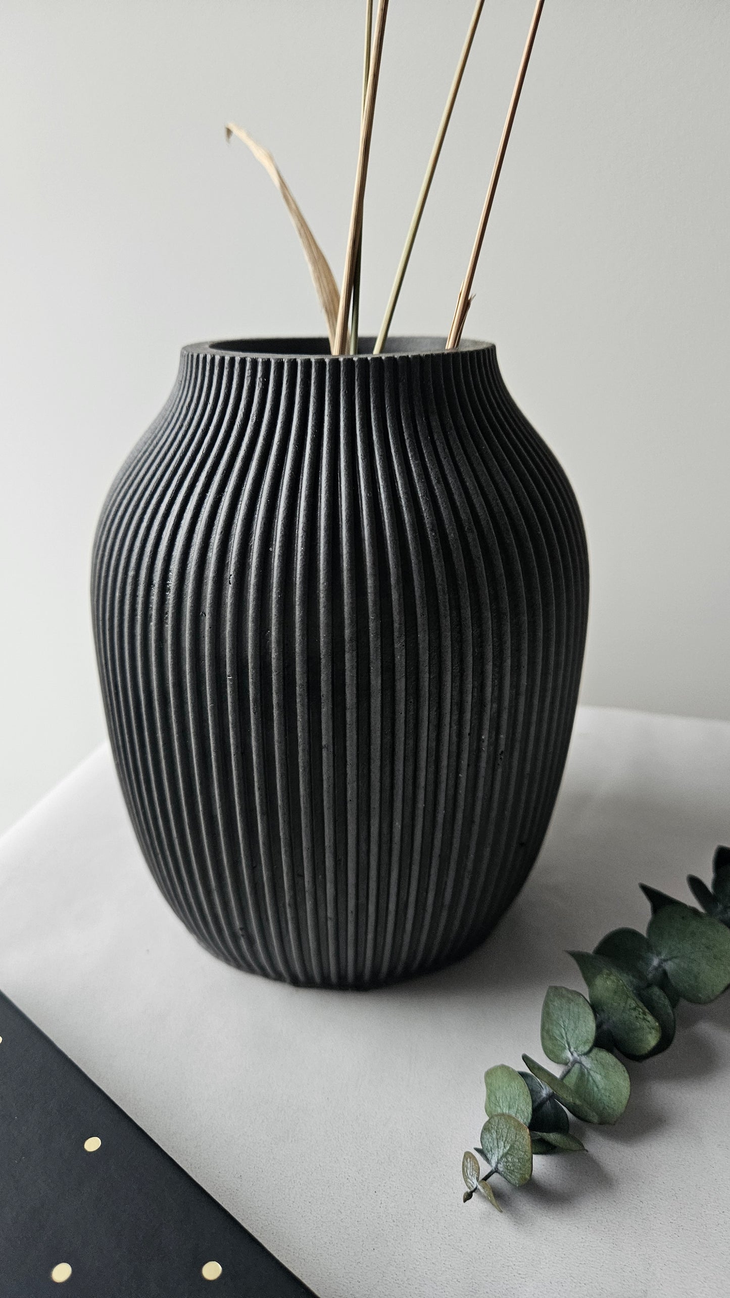 Juoda rumbuota betoninė vaza su auksinėmis šakomis ir žaliais lapais – moderni minimalistinio interjero detalė.