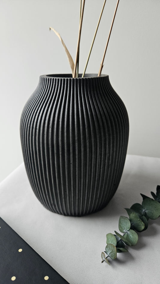 Juoda rumbuota betoninė vaza su auksinėmis šakomis ir žaliais lapais – moderni minimalistinio interjero detalė.