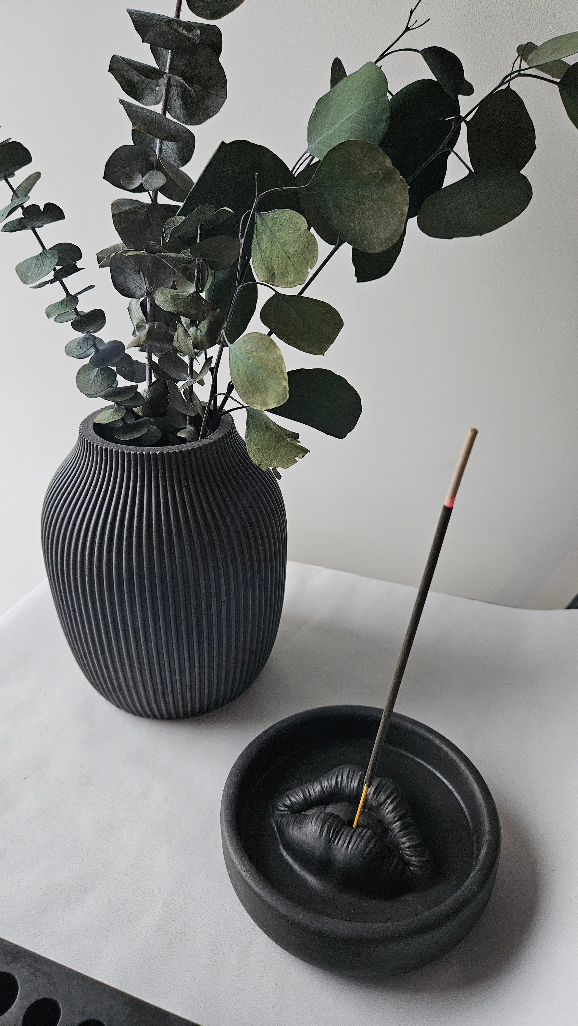 Rankų darbo juoda rumbuota betoninė vaza su auksinėmis šakomis ir žaliais lapais, skirta moderniam ir minimalistiniam interjerui.