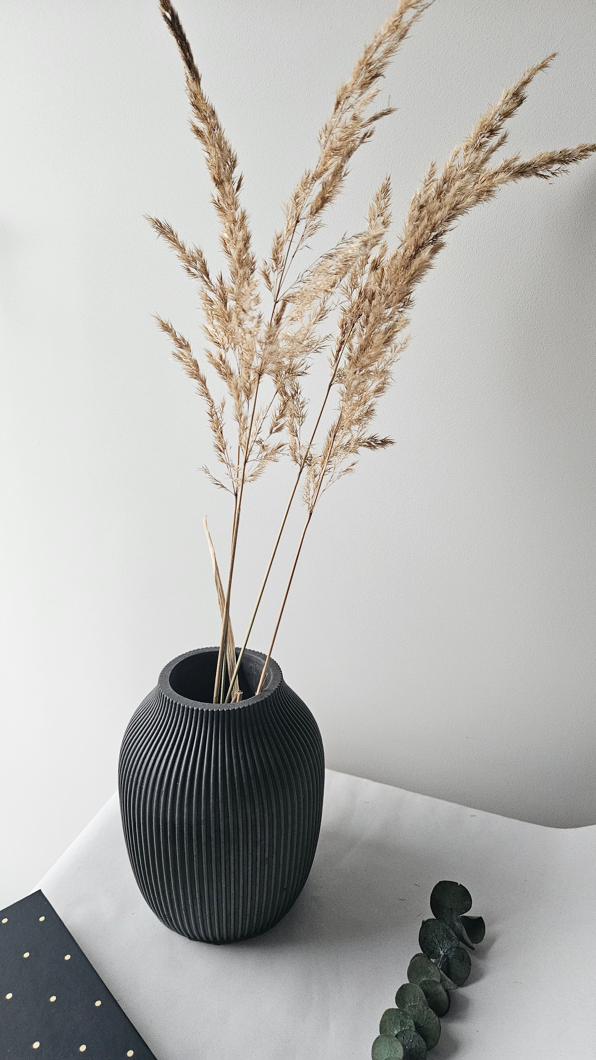 Juoda rumbuota betoninė vaza su auksinėmis šakomis ir žaliais lapais, minimalistinis interjero dekoras.